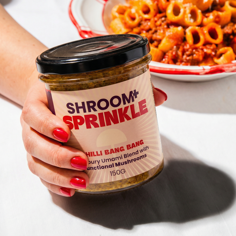Hand holding a jar of 'Shroom+ Sprinkle Chilli Bang Bang' with a bowl of pasta in Napolitana sauce in the background
